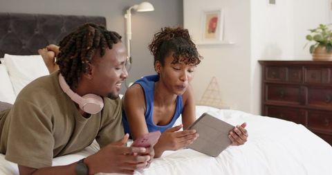 African american couple relaxing on bed sharing tablet and phone, cozy bedroom moment