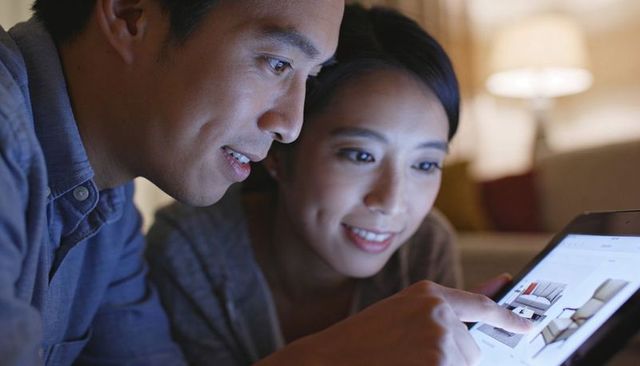 Asian couple browsing furniture on tablet at home, cozy evening lamplight and smiles