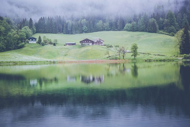 Beautiful serene scene featuring a calm Alpine lake reflecting misty hills, rustic houses, and lush greenery. Perfect for travel brochures, nature blogs, relaxation content, and meditation apps.