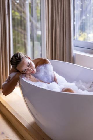 Serene woman reclining in freestanding bathtub with foam and natural light for wellness