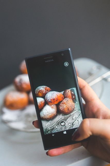 Young person using smartphone to photograph powdered doughnuts on table. Useful for content related to food photography, culinary blogs, bakeries, cooking classes, and social media tutorials.