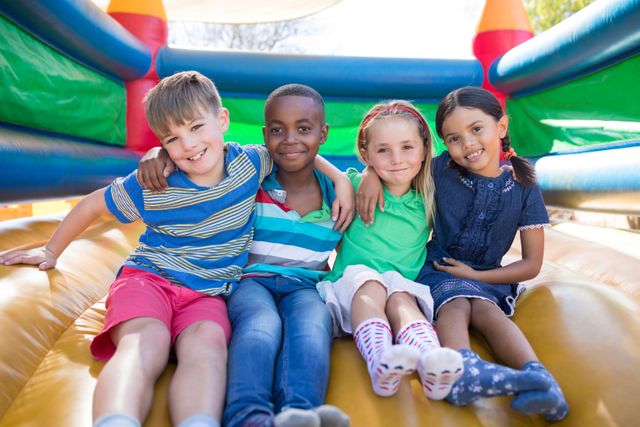This image shows a group of diverse children sitting together on a bouncy castle at a playground. They are smiling and have their arms around each other, indicating strong friendship and joy. This image can be used for promoting children's activities, playgrounds, summer camps, or any content related to childhood fun and bonding.