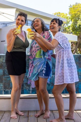 Diverse women laughing and toasting cocktails on sunny deck in swimwear