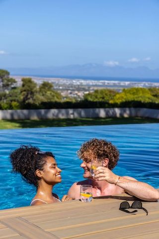 Romantic couple relaxing in infinity pool raising lemon drinks on wooden deck sea view