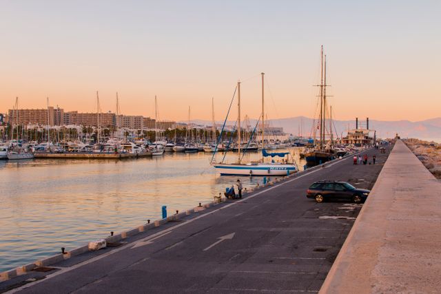 Sunset view of a marina with various sailing boats docked. Calm waters reflect the warm colors of the twilight sky. buildings and mountains are visible in the background. Ideal for depicting travel destinations, vacation landscapes, nautical themes, urban planning, promotional tourism material, or relaxation contexts.