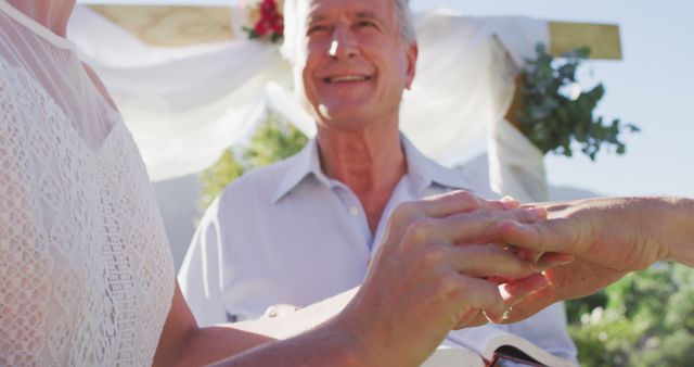 Senior couple exchanging wedding rings during an outdoor ceremony, capturing a joyful and romantic moment. Can be used for topics related to love in later life, weddings, senior romance, marriage, and family celebrations.