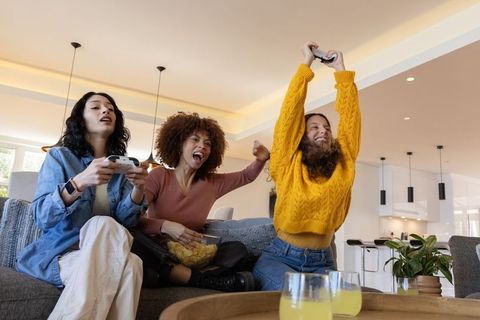 Diverse friends celebrating gaming victory on couch, cheering with controllers and snacks