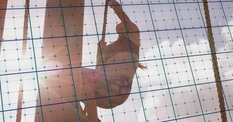 Backlit athlete climbing rope on outdoor obstacle course silhouette behind blue gridlines