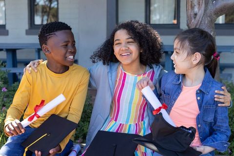 Multicultural elementary graduates smiling on campus bench holding diplomas and caps