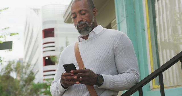 Mature man in casual clothes standing outside, engaged with his smartphone. Modern building and green structure in background. Perfect for themes of technology use, online communication, modern lifestyles, or outdoor casual activities.