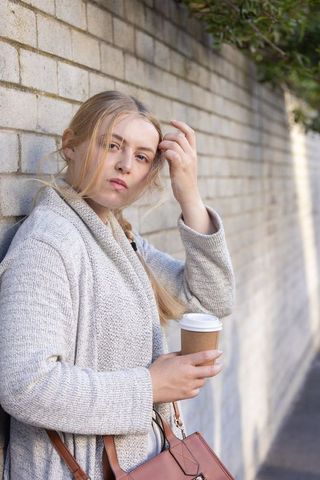 Young woman leaning on brick wall holding takeaway coffee cup with leather handbag