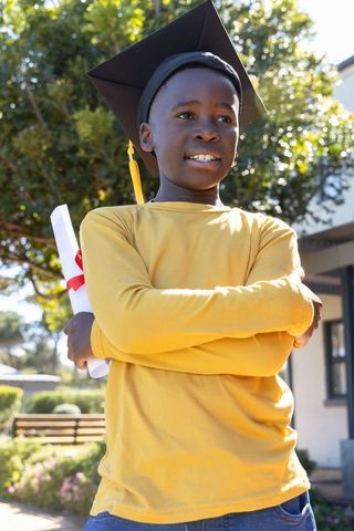 African american child celebrating graduation wearing mortarboard holding diploma outdoors