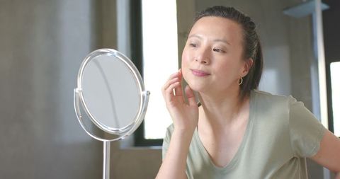 Woman examining skin in mirror during minimal morning skincare routine natural light