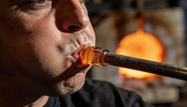 Glassblower shaping molten glass on blowpipe close-up with furnace glow in workshop