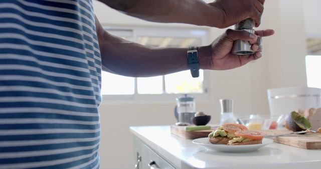 African American man sprinkling seasoning on avocado toast in modern kitchen setting. Ideal for illustrating themes of healthy eating, home cooking, culinary skills, breakfast preparation, and domestic lifestyle. Suitable for food blogs, cooking articles, recipe websites, and lifestyle content.