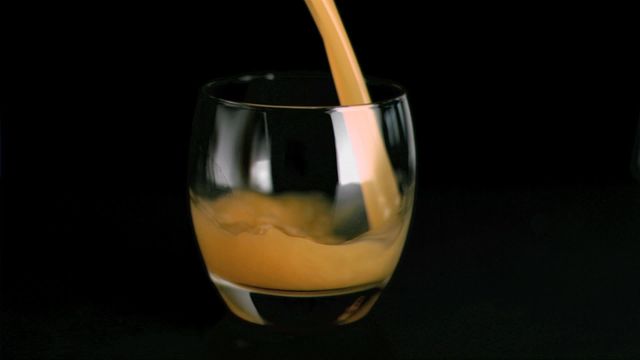 Fruit juice pours into a clear glass in super slow motion, set against a stark black background. The smooth flow and vibrant color of the juice create a visually appealing contrast. Ideal for advertising beverages, highlighting health benefits of fruit juices, use in food blogs, or illustrative purposes in nutrition presentations.