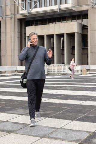 Middle-aged man walking and talking on phone in urban plaza with gym bag and water bottle