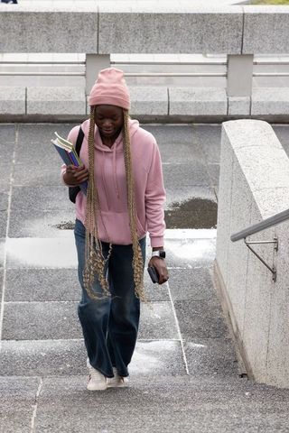 Adult african american woman walking up stairs in pink beanie and hoodie carrying notebooks