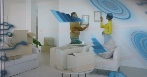 Couple hanging wall art in modern living room amid moving boxes and tech overlays