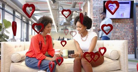Two coworkers chatting on office sofa holding coffee and device surrounded by red hearts