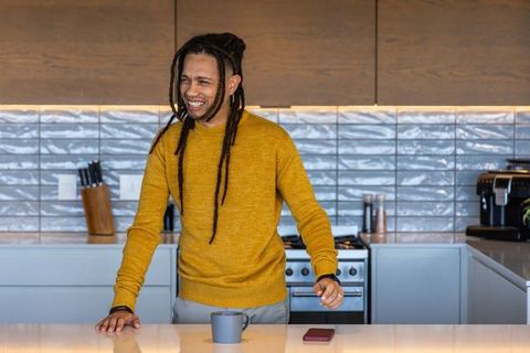 Smiling dreadlocked man leaning on kitchen island wearing mustard sweater with mug and phone