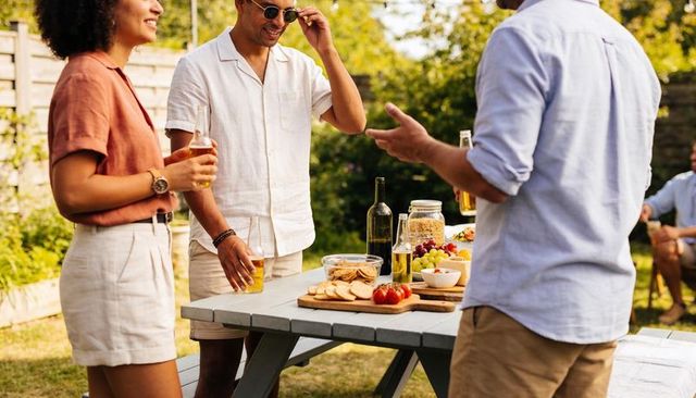 Friends chatting and enjoying drinks at gray picnic table in sunny backyard