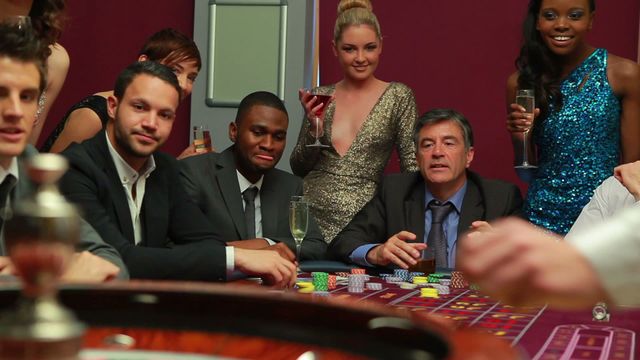 Sophisticated group in formal attire gathers around roulette table at lavish casino, engrossed in the thrill of gambling. Atmosphere filled with anticipation and fine drinks. Ideal for themes related to luxury entertainment, nightlife adventures, or high-stakes wagering excitements. Perfect for advertising recreation and leisure experiences inclusive of style and excitement.