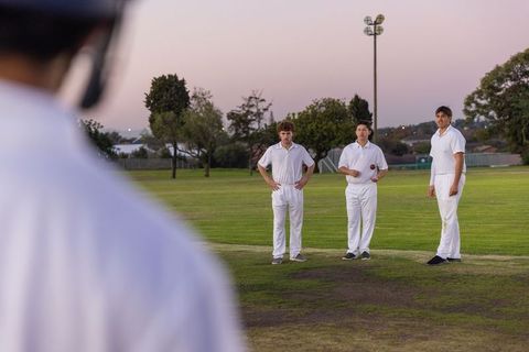 Three cricket players standing on worn dusk pitch in white uniforms holding red ball