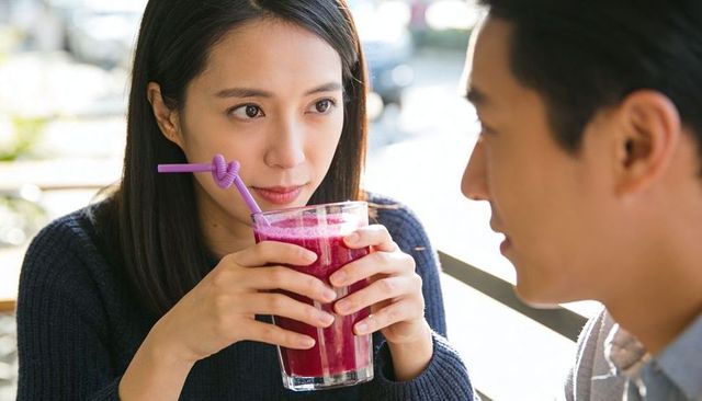Asian woman sipping vibrant magenta smoothie with purple straw while listening to partner