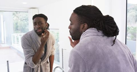 African American man examining skin in bathroom mirror wearing bathrobe with dreadlocks