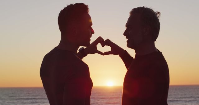 Happy caucasian gay male couple holding hands and making heart shape at sunset on the beach. summer road trip and holiday in nature.