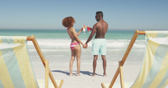 Couple standing on sandy beach, holding hands, and enjoying the ocean view. Ideal for use in travel promotions, romantic getaway advertisements, tropical vacation packages, or leisure lifestyle content.