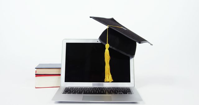 Image shows a graduation cap resting on a laptop screen with textbooks beside. Perfect for use in online education materials, academic achievement posts, e-learning promotions, or distant learning brochures. Represents modern education and the merging of technology with learning.