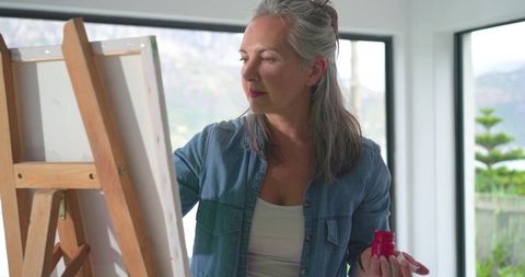 Mature woman painting on canvas at easel in bright sunlit home studio