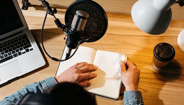 Podcaster writing notes in home studio with microphone, laptop and coffee cup