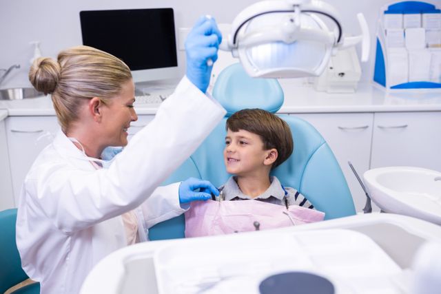 Dentist adjusting light while young boy sits in dental chair, smiling. Ideal for use in healthcare, pediatric dentistry, dental care promotions, and medical professional training materials.