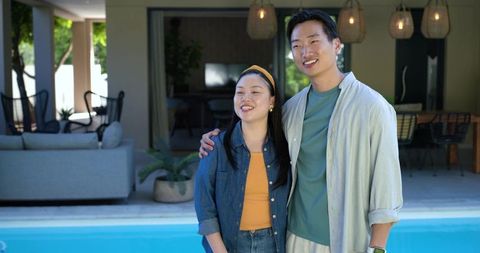 Smiling asian couple standing by modern poolside patio enjoying sunny outdoor lounge