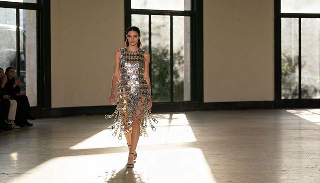 Model walking runway wearing reflective metal disc dress catching sunlight and shadows
