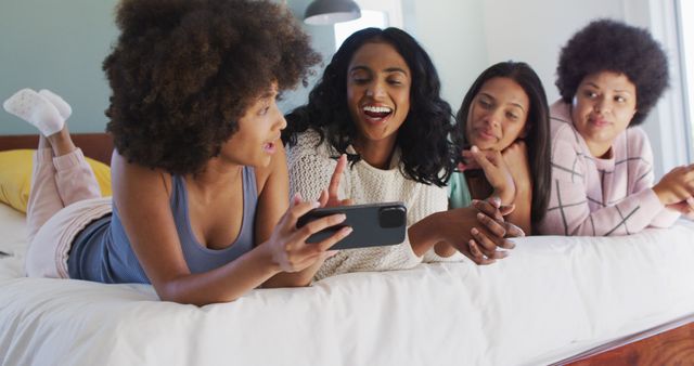 Happy diverse female friends lying on bed and using smartphone in bedroom. spending quality time at home.