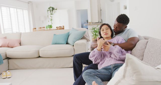 Interracial couple sitting on couch relaxing with hot drinks in modern living room during daytime. Suitable for themes on relationships, leisure time, home comfort, and interracial love.
