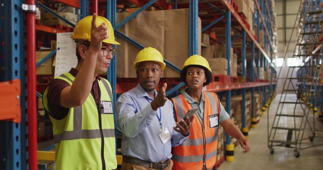 Three warehouse managers are coordinating and discussing inventory operations in a large storage facility. They are wearing safety vests and hard hats for protection. This image can be used in articles or websites related to logistics, warehouse management, industrial operations, workplace safety, or managing inventory.