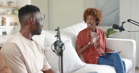 Podcast cohosts recording candid interview on living room sofa with professional mics