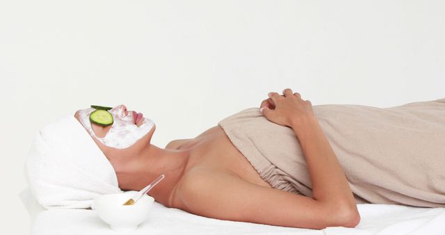 A young Caucasian woman enjoys a relaxing facial treatment at a spa, with copy space. Her face is covered with a mask and cucumber slices over her eyes, symbolizing self-care and beauty routines.