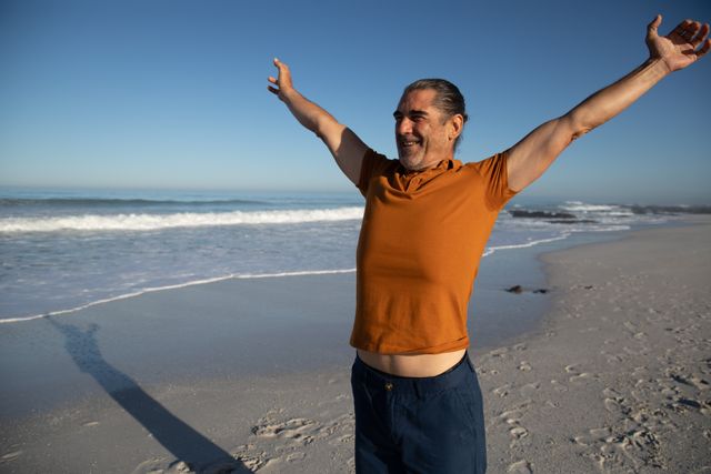 This image captures a senior Caucasian man enjoying a sunny day at the beach with his arms outstretched, symbolizing freedom and happiness. The ocean waves and clear blue sky create a serene and relaxing atmosphere. Ideal for use in advertisements, travel brochures, retirement planning materials, and wellness campaigns.