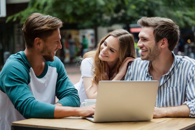 Young couple enjoying time with friend at a sidewalk cafe in the city. They are smiling and chatting while using a laptop. Perfect for themes related to friendship, urban lifestyle, leisure activities, and social interactions.