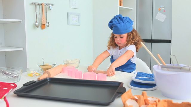 A young girl in a blue chef hat focuses on baking heart-shaped cookies on a kitchen counter. Various baking tools like mixing bowls, cookie cutters, and baking sheets surround her. This image beautifully captures a moment of childhood fun and cooking creativity, ideal for illustrating topics such as children’s baking activities, family recipes, or kitchen safety for kids.