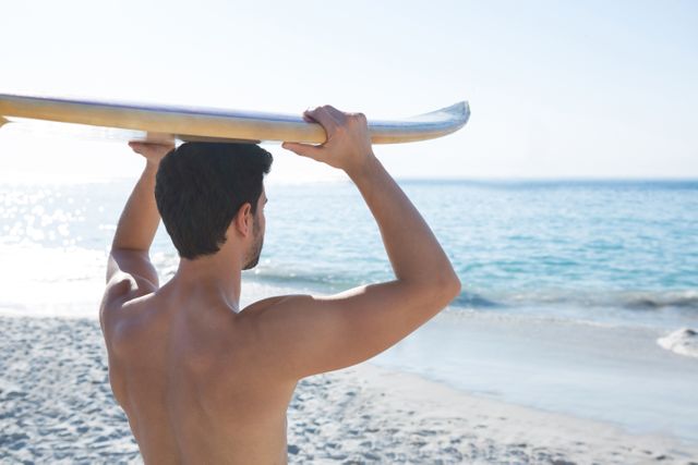 Rear view of shirtless man carrying surfboard at beach on sunny day