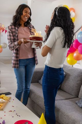 Mother presenting lit birthday cake to teenage daughter during colorful home celebration