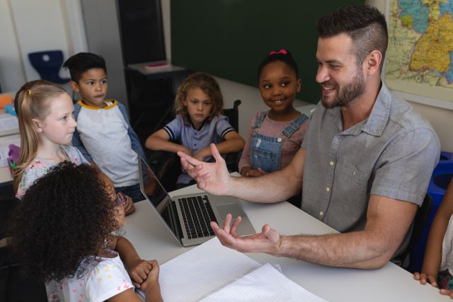 Male teacher engaging with a group of diverse elementary school students in a classroom. The children are gathered around a desk, attentively listening and interacting with the teacher who is using a laptop. Ideal for educational content, school brochures, teacher training materials, and articles on classroom dynamics and primary education.