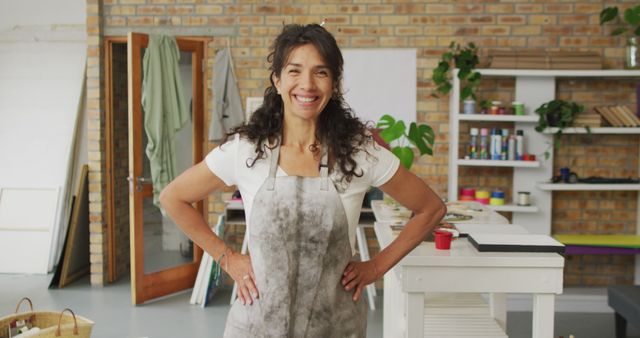 Woman standing confidently in an art studio, wearing an apron. Background features art supplies, shelves, and tools for creative work. Ideal for use in advertising creative professions, promoting art classes, or showcasing artist profiles.